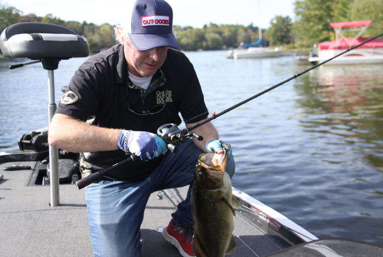 An angler proudly holds a large largemouth bass aboard a bass boat, with the lake stretching out behind them—a testament to the effectiveness of the Bait Finesse System, which offers anglers a fresh approach to targeting bass with light-action baitcasting gear and precise lure selection.